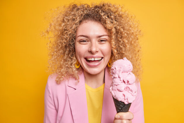 糖一位留着卷发的漂亮女人的肖像她拿着锥形冰激凌 在夏天和朋友们在公园里散步 被隔离在黄色的墙壁上女模特吃着美味的甜点室内夹克味道
