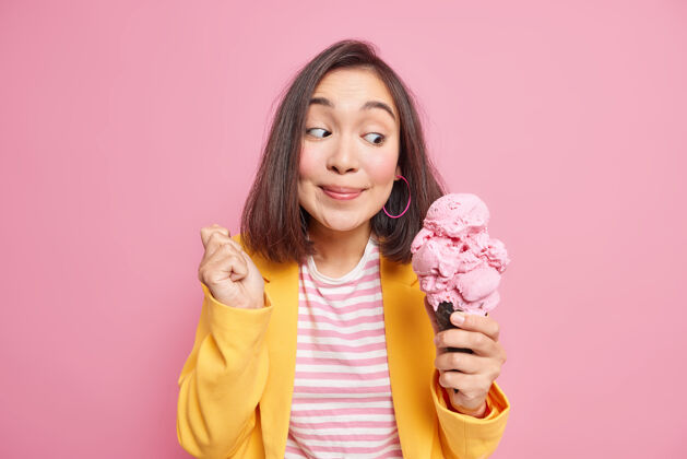 服装快乐惊讶的亚洲年轻女子看着开胃的大冰淇淋微笑愉快地享受着吃的东西美味的休息饮食穿着时髦的衣服隔离在粉红色的墙上个人东方小吃