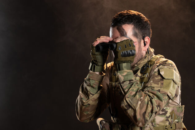伪装身穿军装的男战士透过望远镜在黑暗的墙上看军队战争衣服