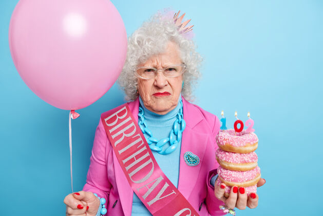 生日不满的老太太横拍皱眉脸上有不开心的心情拿着一堆美味的油炸圈饼和燃烧的蜡烛充气气球唇膏人珠宝