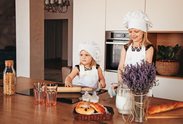 女孩厨房里两个滑稽的女孩戴着厨师帽和白色围裙在厨房里玩金发面包师幼儿