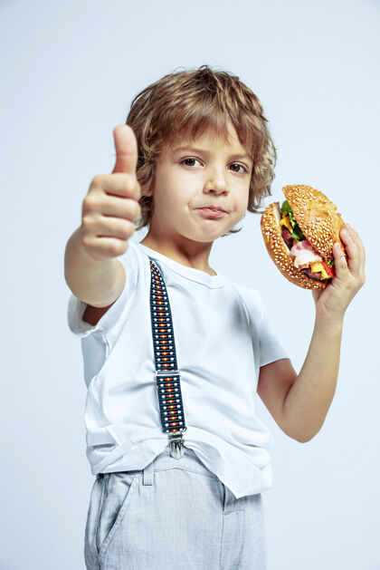 肖像在白色工作室墙上穿着休闲服的漂亮小卷发男孩正在吃汉堡情感人欢呼
