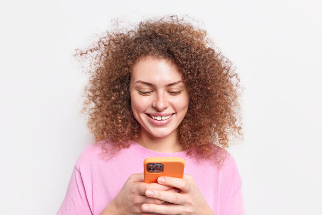 乐观积极的卷发年轻的欧洲女人微笑着轻轻地拿着现代智能手机在社交媒体上聊天连接到无线互联网穿着休闲服隔着白色的墙壁使用应用程序微笑卷发电话