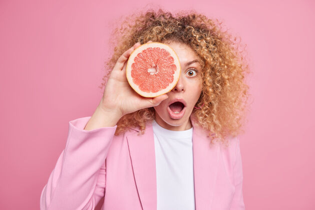 卷发惊讶的卷发欧洲女人用葡萄柚片遮住眼睛 穿着时尚的衣服张开嘴 隔着粉色的墙壁夏日茶点柑橘类水果概念罗西水果果汁