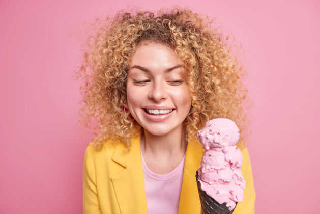 风味室内拍摄的漂亮开朗的欧洲女人开心地微笑着看着开胃的粉红色冰淇淋在黑色华夫饼穿着黄色夹克自然卷发隔离在玫瑰色的墙壁惊人欢乐欧式
