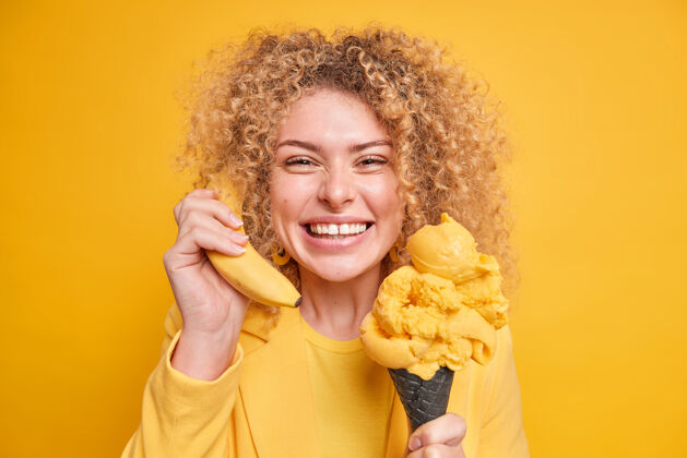 奶油积极的卷发女人玩得很开心喜欢吃柠檬味的美味冰淇淋把香蕉放在耳边假装打电话给别人表达积极的情绪隔着黄色的墙营养牙齿香蕉