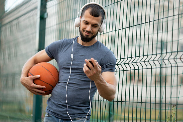 运动员街上一个男人手里拿着篮球 看着智能手机 戴着耳机听音乐人力量健身