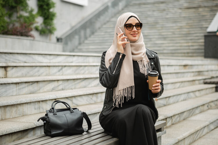 手机现代时尚的妇女戴着头巾 皮夹克和黑色长袍 戴着太阳镜坐在城市街道上讲手机文化传统迪拜