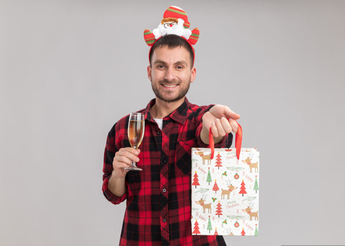 年轻微笑着的白人年轻人戴着圣诞头带 手里拿着一杯香槟 面带微笑地向隔离在白色墙壁上的相机伸出圣诞礼品袋 并留有复印空间男人穿头带