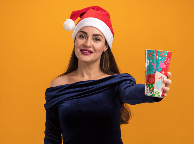 杯子微笑着的年轻漂亮的女孩穿着蓝色的裙子 戴着圣诞帽 举着圣诞杯孤立在橙色的墙上年轻衣服帽子
