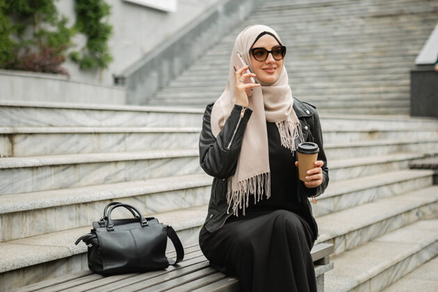手机现代时尚的穆斯林妇女戴着头巾 皮夹克和黑色长袍 戴着太阳镜坐在城市街道上讲手机文化传统迪拜
