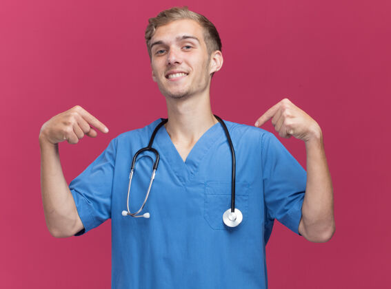 公民自信的年轻男医生穿着医生制服 手持听诊器 指着隔离在粉红色墙上的自己衣服听诊器医生
