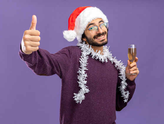 微笑微笑的年轻帅哥戴着圣诞帽 脖子上戴着花环 手里拿着一杯香槟 在蓝色的墙上孤立地竖起大拇指小伙子帽子玻璃杯