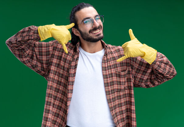 男人带着微笑的年轻帅气的清洁工 穿着t恤 戴着手套 在绿色的墙上显示着电话的手势人衣服表演
