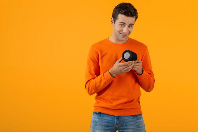 时髦时尚微笑的年轻人穿着橙色毛衣手持无线扬声器快乐地听音乐玩得开心音乐娱乐家伙
