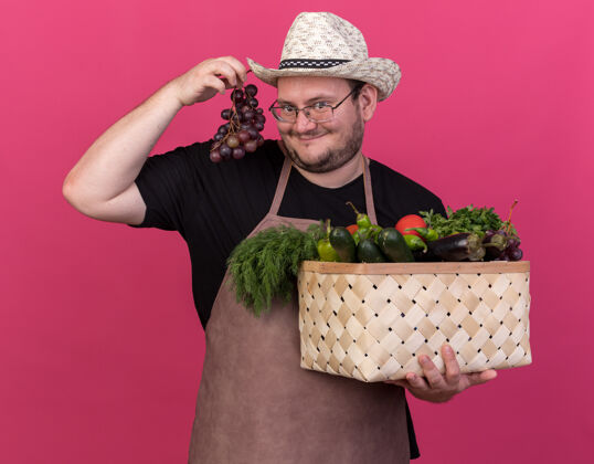 蔬菜高兴的年轻男园丁戴着园艺帽 手里拿着菜篮子 把葡萄隔离在粉红色的墙上姿势葡萄感觉