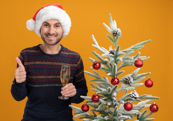男人微笑着的白人年轻人戴着圣诞帽 站在装饰好的圣诞树旁 手里拿着一杯香槟 看上去孤立地站在橙色的墙上 竖起大拇指香槟圣诞节高加索