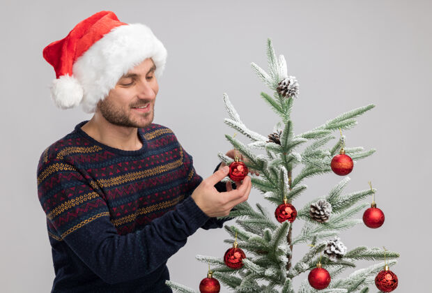 圣诞节微笑着戴着圣诞帽的白人年轻人站在圣诞树旁看着它触摸着隔离在白色墙上的圣诞饰品年轻人白种人微笑