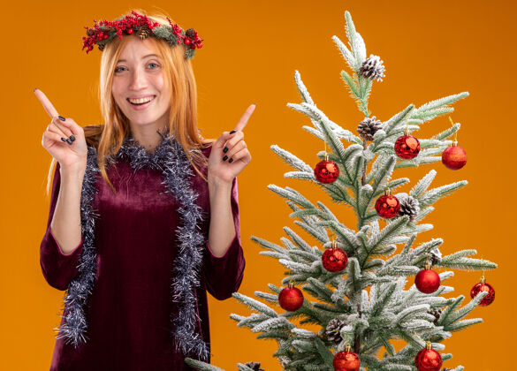 年轻微笑着站在圣诞树旁的年轻漂亮的女孩穿着红色的裙子 脖子上戴着花环 点在橙色的墙上衣服站穿