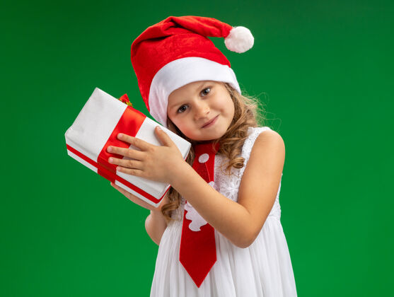 小高兴的歪头小女孩戴着圣诞帽打着领带拿着礼品盒隔离在绿色的墙上女孩请帽子