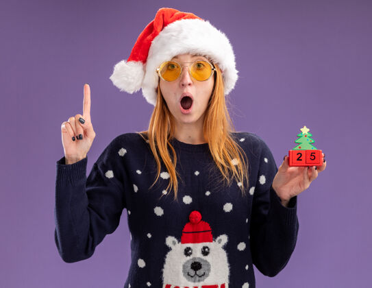 穿惊讶的年轻漂亮女孩穿着圣诞毛衣 戴着圣诞帽 戴着圣诞眼镜 拿着圣诞玩具点着 孤零零地站在紫色的墙上毛衣眼镜举行