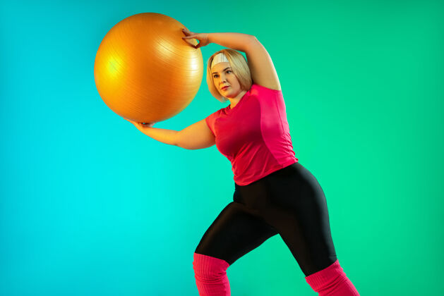 训练年轻的白人大号女模特在霓虹灯下的渐变绿色背景下训练用健身球做运动运动的概念 健康的生活方式 积极的身体 平等轻积极霓虹灯