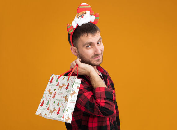 圣诞老人戴着圣诞老人头带的年轻白种人站在侧视图中 肩上扛着圣诞礼品袋 看起来孤立地站在橙色的墙上 留着复制空间男人穿立场