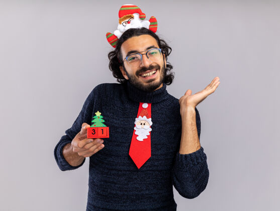 手笑容可掬的年轻帅哥戴着圣诞领带 戴着发箍 手拿着圣诞玩具点 隔离在白色墙壁上 留着复制空间帅哥圣诞节年轻