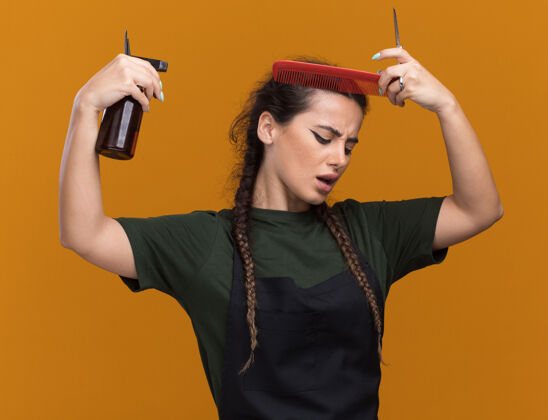 女性年轻的女理发师穿着制服 梳头发 用喷雾瓶在橙色的墙上隔离洒水头公民姿势制服
