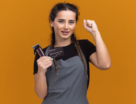 头发高兴的年轻女理发师穿着制服拿着信用卡和理发器显示是的手势隔离在橙色的墙上剪子秀是