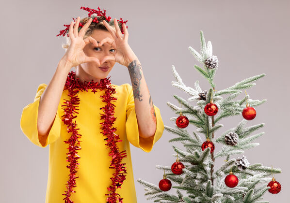 女人自信的金发女郎 头戴圣诞花环 脖子上戴着金属丝花环 站在装饰好的圣诞树旁 在隔离在白墙上的脸前做着心形手势脸头脖子
