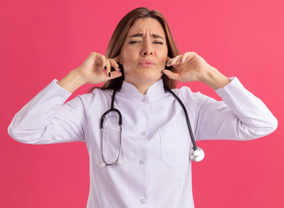 人穿着医用长袍 戴着听诊器的年轻女医生 耳朵被隔离在粉红色的墙上脸衣服站立