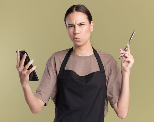 制服困惑的年轻黑发理发师女孩穿着制服拿着头发稀疏剪刀和电话困惑绿色理发师