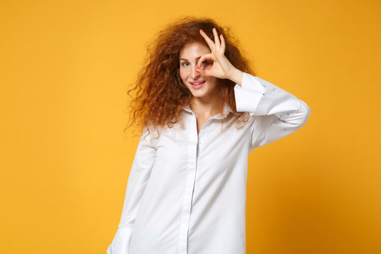 时尚穿着休闲白衬衫的年轻红发女郎在黄橙色的墙上孤零零地摆姿势一个成人站立