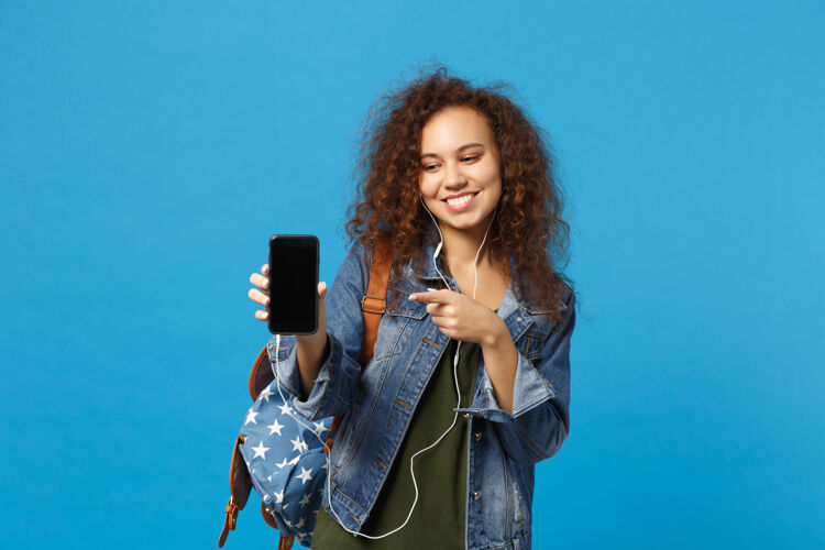 音频年轻的非洲裔美国少女 穿着牛仔服 背着耳机 隔离在蓝色的墙上耳机屏幕女性