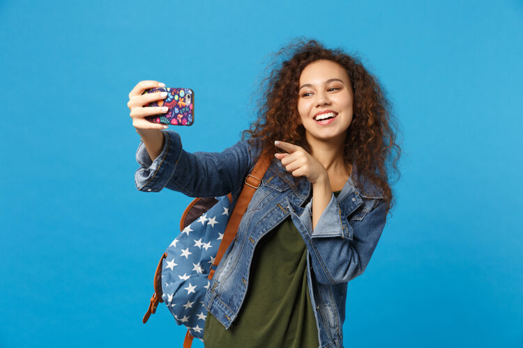 电话年轻的非洲裔美国女孩十几岁的学生在牛仔服装 背包举行电话隔离在蓝色的墙壁上包女人手机