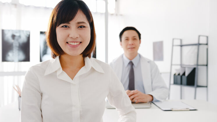 人自信的亚洲男医生穿着白色的医疗制服 年轻的女病人看着相机 微笑着在诊所或医院的服务台上接受医疗咨询制服教授微笑