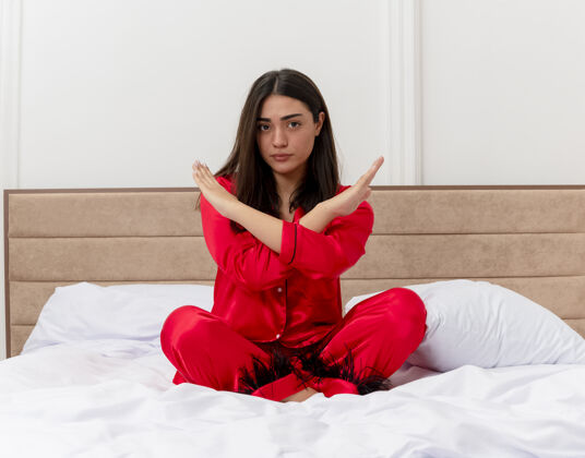 脸穿着红色睡衣的年轻美女坐在床上做着停下来的手势 在卧室里严肃地交叉着双手交叉漂亮红