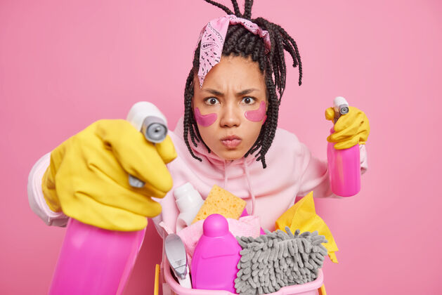 手套男人留着长发锁 摆着带清洁用品的姿势 戴着橡胶手套 拿着清洁剂喷雾剂或隔离在粉红色上的洗涤剂穿家庭主妇女人