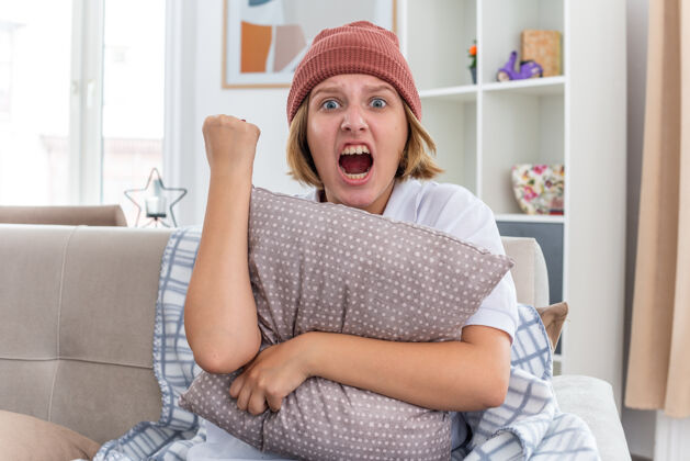 表情愤怒的不健康的年轻女子 戴着暖和的帽子 拿着毯子 看起来不舒服 患感冒和流感 抱着枕头 握紧拳头 带着咄咄逼人的表情 坐在客厅的沙发上流感生气年轻