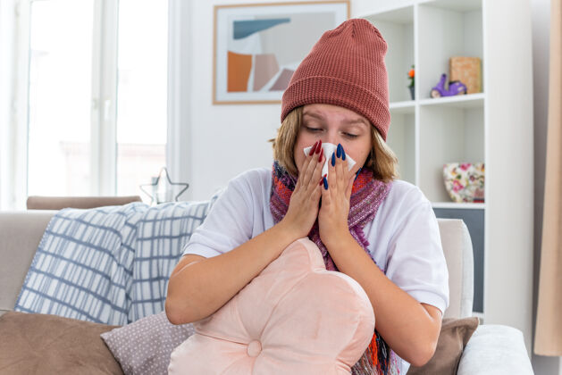 光不健康的年轻女子戴着暖和的帽子 戴着围巾 抱着枕头 用纸巾擤鼻涕 患有感冒和流感 坐在明亮的客厅椅子上流感年轻打击