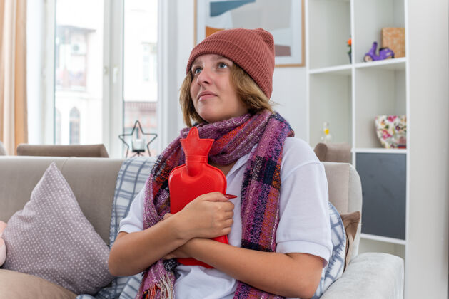 女人不健康的年轻女子戴着帽子 脖子上围着暖和的围巾 感觉不舒服 生病 患感冒和流感 拿着热水瓶 看起来很担心 坐在客厅里的沙发上脖子冷不适