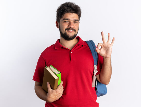 人年轻的留着胡子的学生 穿着红色马球衫 背着背包 拿着笔记本 微笑着 白墙上挂着“ok”的牌子笔记本背包马球