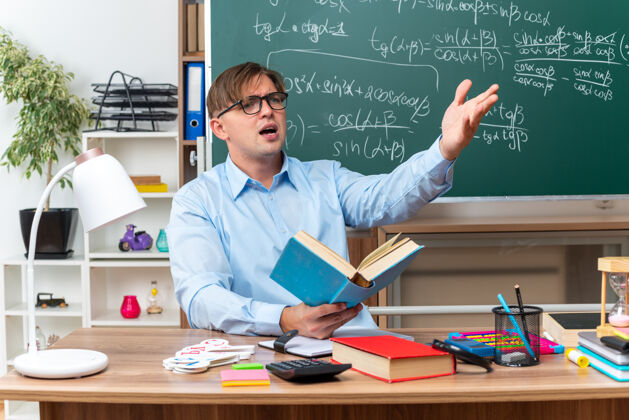 学校戴着眼镜的年轻男老师困惑而不快地站在一边愤怒地举起手臂坐在课桌旁 教室里的黑板前放着书和笔记困惑愤怒一旁