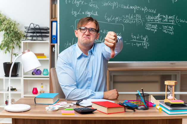 教室戴眼镜的年轻男老师不高兴地竖起大拇指坐在课桌旁 教室的黑板前放着书和笔记拇指学校课桌