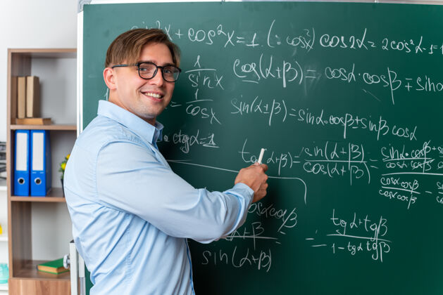 公式戴着眼镜的年轻男老师在教室里兴高采烈地微笑着站在黑板旁讲解数学公式穿教室解释