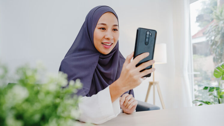 工作年轻的穆斯林女商人使用智能手机通过视频聊天与朋友交谈头脑风暴在线会议 同时在客厅远程工作网络团队青少年