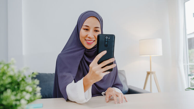 工作年轻的女商人使用智能手机通过视频聊天与朋友交谈头脑风暴在线会议 同时在客厅远程工作办公室头巾虚拟