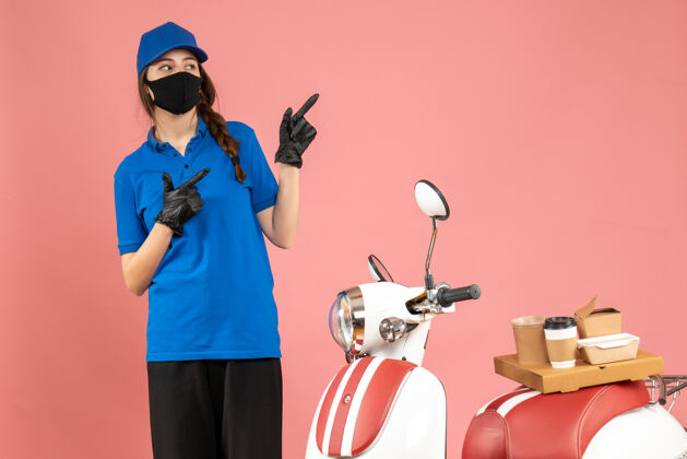 咖啡俯视图：戴着医用口罩的信使女孩站在摩托车旁边 咖啡蛋糕在柔和的桃色背景上指向上人物粉彩面具