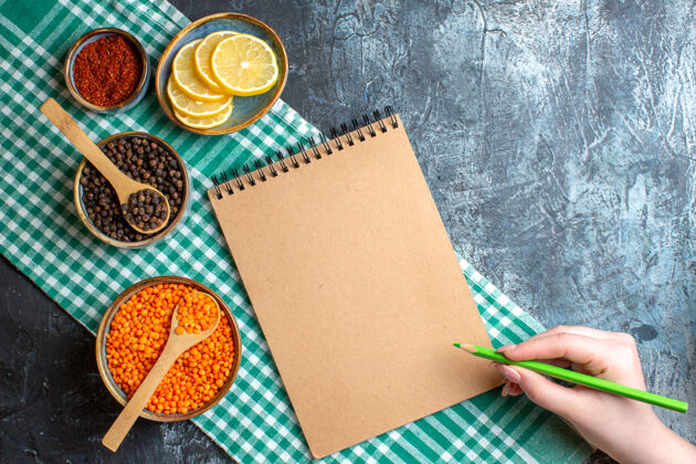 拿着上图为晚餐背景 用不同的香料黄豌豆和手拿笔放在黑色桌上的螺旋笔记本上包装深色信封